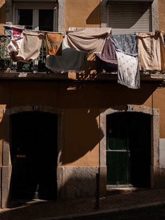 Laundry hangs from a balcony railing, casting shadows on the yellow wall of a building. Various fabrics and towels of different colors and patterns are draped over the railing. Below, two dark green doors provide a contrast to the sunlit textiles above.