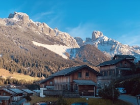 Rustic wooden houses are nestled in a valley with majestic snow-capped mountains in the background. The clear blue sky complements the serene landscape, while dense forests wrap around the base of the mountains.