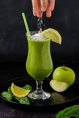 A green smoothie in a tall, elegant glass garnished with a lemon slice on the rim. A hand is sprinkling grated coconut or another white powder onto the smoothie, and a green straw is placed in the drink. Around the glass are fresh spinach leaves, lemon slices, and a green apple, one half slightly cut. Everything is set against a dark background.