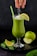 A green smoothie in a tall, elegant glass garnished with a lemon slice on the rim. A hand is sprinkling grated coconut or another white powder onto the smoothie, and a green straw is placed in the drink. Around the glass are fresh spinach leaves, lemon slices, and a green apple, one half slightly cut. Everything is set against a dark background.