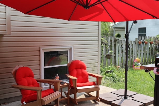 Two red-cushioned wooden chairs are placed under a large red umbrella in a backyard setting. A side table is positioned between the chairs, and there is a window on the beige siding of the house. A wooden fence with hanging flower pots is visible in the background, along with a lawn and a grill.