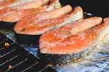 Several slices of salmon are being grilled on a metal rack with aluminum foil underneath. The salmon has a pinkish-orange color with visible black pepper seasoning on top.