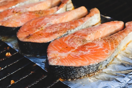 Several slices of salmon are being grilled on a metal rack with aluminum foil underneath. The salmon has a pinkish-orange color with visible black pepper seasoning on top.