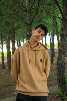 A young individual wearing a tan hoodie standing amidst a forest of tall trees with dense green foliage. The person has a relaxed posture with a slight tilt of the head and hands behind the back. The background features rows of trees and sunlight filtering through the leaves, creating a serene and natural atmosphere.