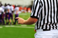 A referee wearing a black and white striped uniform points towards a football field. In the background, there are blurred players wearing purple uniforms, suggesting a sporting event is taking place.