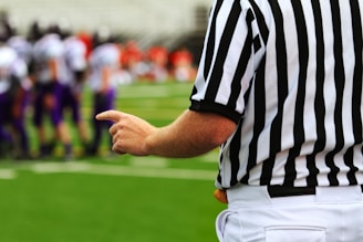 A referee wearing a black and white striped uniform points towards a football field. In the background, there are blurred players wearing purple uniforms, suggesting a sporting event is taking place.