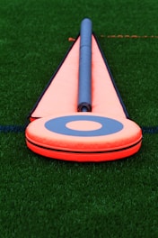 A bright orange football field marker with a blue circle in the center lies on the green turf. The marker is triangular with a cylindrical rod pointing outward.