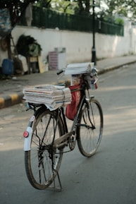 A bicycle is parked on a street, loaded with a stack of newspapers secured on the back and a red bag hanging from the frame. The background includes a blurred view of a sidewalk, a fence, and various objects.