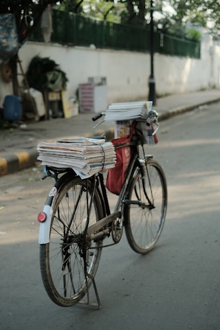 A bicycle is parked on a street, loaded with a stack of newspapers secured on the back and a red bag hanging from the frame. The background includes a blurred view of a sidewalk, a fence, and various objects.