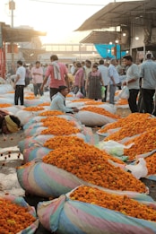 A bustling flower market with numerous sacks filled with vibrant orange marigolds. Many people are gathered around, some standing and others examining or buying flowers. The setting appears outdoors, with a rustic roof structure providing partial cover. The scene is lively, capturing the essence of a busy marketplace.