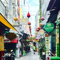 A narrow alleyway adorned with colorful hanging lanterns and vibrant signage from various businesses, including restaurants and hotels. Scooters are parked on one side, while a person walks down the street, creating a lively and bustling atmosphere.