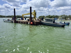 A barge with several people is on water, equipped with a crane and surrounded by various small boats. The sky is partly cloudy, and the water is a light green color. Trees line the distant shore, creating a scenic backdrop.