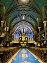 A majestic interior of a church with an ornate ceiling featuring intricate patterns and arches. The sanctuary is illuminated with a soft glow highlighting the blue and gold accents. Rows of wooden pews lead towards a grand altar with decorative columns and stained glass.