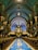 A majestic interior of a church with an ornate ceiling featuring intricate patterns and arches. The sanctuary is illuminated with a soft glow highlighting the blue and gold accents. Rows of wooden pews lead towards a grand altar with decorative columns and stained glass.