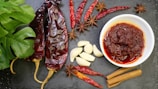 A variety of ingredients are artfully arranged on a dark slate surface. There are dried red chili peppers, garlic cloves, star anise, and cinnamon sticks surrounding a white bowl filled with a chunky red sauce. A lush green leaf adds a splash of color on the left side.