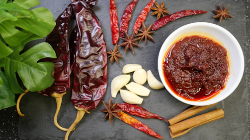 A variety of ingredients are artfully arranged on a dark slate surface. There are dried red chili peppers, garlic cloves, star anise, and cinnamon sticks surrounding a white bowl filled with a chunky red sauce. A lush green leaf adds a splash of color on the left side.