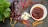 A variety of ingredients are artfully arranged on a dark slate surface. There are dried red chili peppers, garlic cloves, star anise, and cinnamon sticks surrounding a white bowl filled with a chunky red sauce. A lush green leaf adds a splash of color on the left side.
