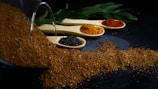 A pile of brown spice has spilled from a glass container and is spread across a dark surface. Three wooden spoons containing various spices are lined up behind the pile. One spoon has black mustard seeds, another contains an orange spice, and the last holds a red spice. Green leaves are visible in the background.