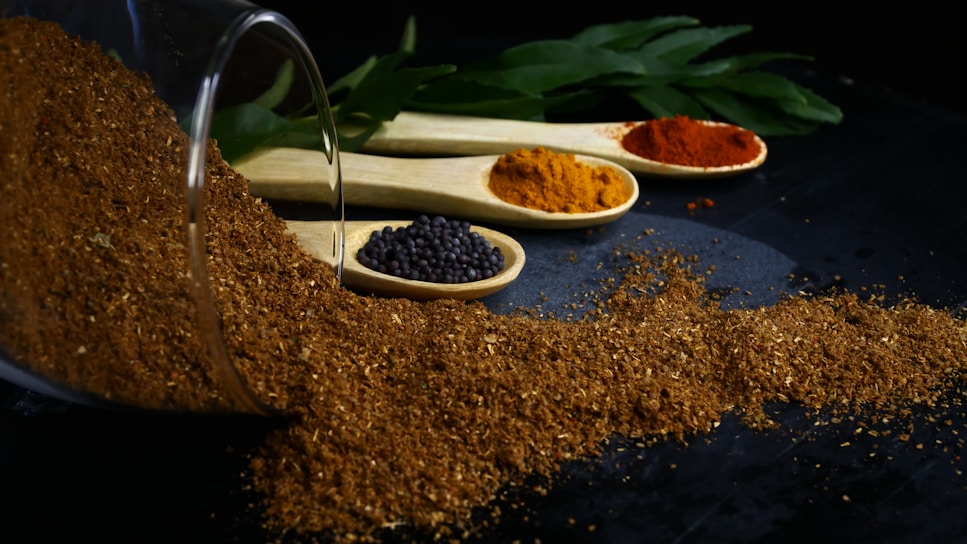 A pile of brown spice has spilled from a glass container and is spread across a dark surface. Three wooden spoons containing various spices are lined up behind the pile. One spoon has black mustard seeds, another contains an orange spice, and the last holds a red spice. Green leaves are visible in the background.
