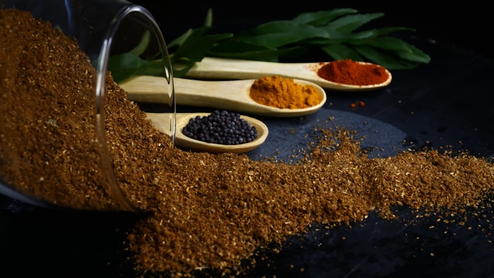 A pile of brown spice has spilled from a glass container and is spread across a dark surface. Three wooden spoons containing various spices are lined up behind the pile. One spoon has black mustard seeds, another contains an orange spice, and the last holds a red spice. Green leaves are visible in the background.