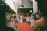A group of women dressed in colorful, floral dresses and wearing decorative headpieces are performing on a red carpet outside a building. The setting appears to be an outdoor event with multi-colored triangular flags hanging overhead. There are other people and vendors in the background, along with some palm trees and city buildings.