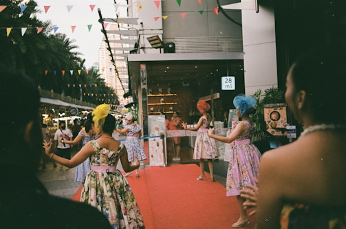 A group of women dressed in colorful, floral dresses and wearing decorative headpieces are performing on a red carpet outside a building. The setting appears to be an outdoor event with multi-colored triangular flags hanging overhead. There are other people and vendors in the background, along with some palm trees and city buildings.