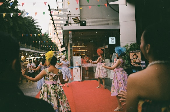 A group of women dressed in colorful, floral dresses and wearing decorative headpieces are performing on a red carpet outside a building. The setting appears to be an outdoor event with multi-colored triangular flags hanging overhead. There are other people and vendors in the background, along with some palm trees and city buildings.