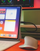 A tablet with a keyboard cover on a desk, showing a screen with app widgets and icons. The device is connected to a cable, and a wireless mouse is placed nearby. A monitor in the background displays colorful graphics.