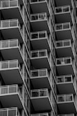 A series of repeating balconies on a tall building, each featuring metal railings and glass panels. The architectural design creates a geometric pattern with strong, clean lines and a sense of uniformity.