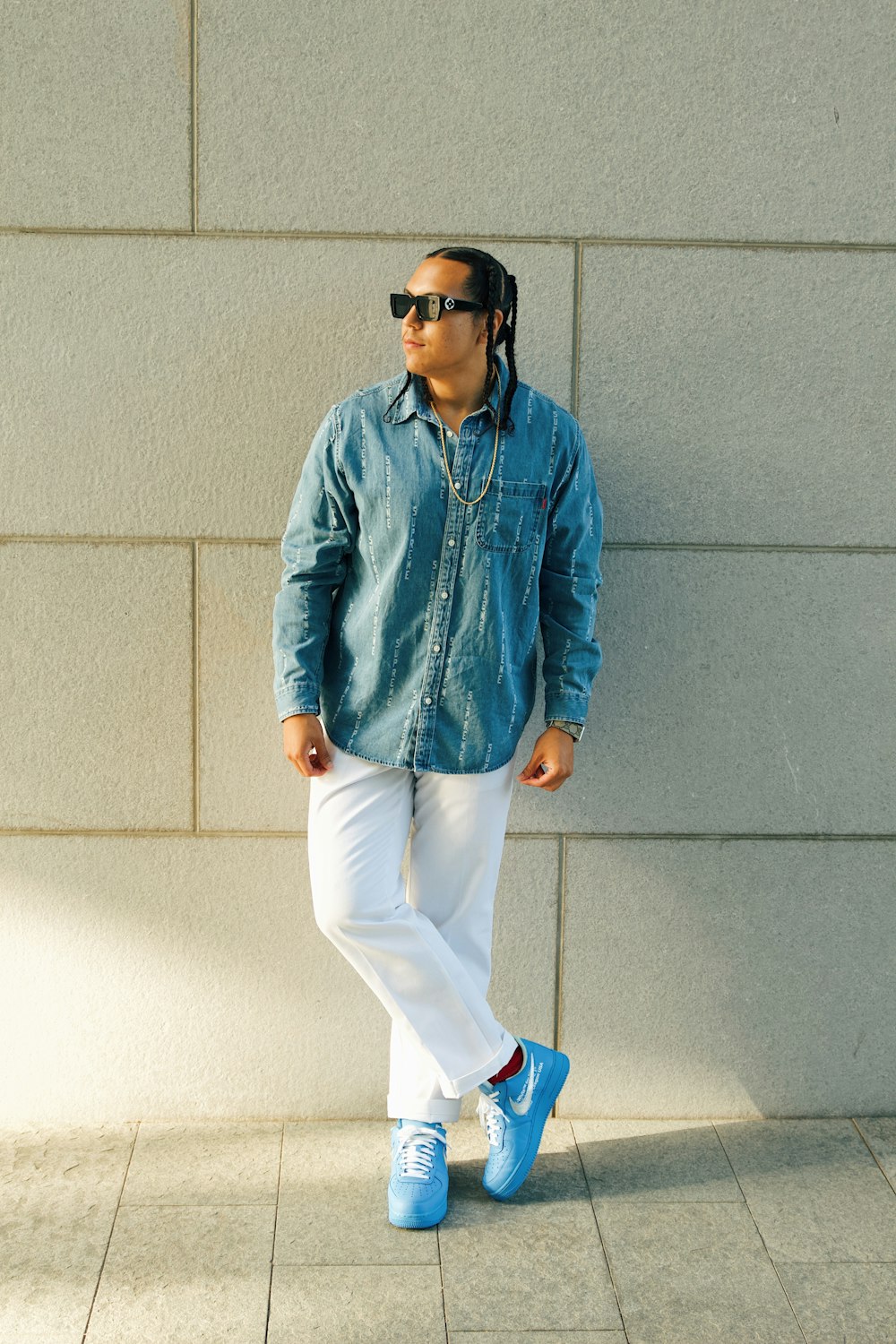 a man standing against a wall wearing sunglasses and a denim shirt