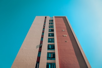 A tall building with a modern design is seen from a low angle, extending upwards towards the bright, clear blue sky. The facade features vertical rows of windows and large letters on a prominent red panel.