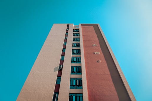 A tall building with a modern design is seen from a low angle, extending upwards towards the bright, clear blue sky. The facade features vertical rows of windows and large letters on a prominent red panel.