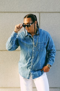 A person stands against a concrete wall, wearing a blue denim shirt with white text, white pants, and sunglasses. The hair is styled in braids, and a gold chain is visible around the neck. The individual exudes a confident and stylish persona, holding the sunglasses with one hand.