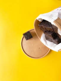 A round cardboard container lies on a vibrant yellow background. Dark brown pieces of chocolate cake or brownie are partially wrapped in white paper, with one piece placed separately on top of the container's lid.