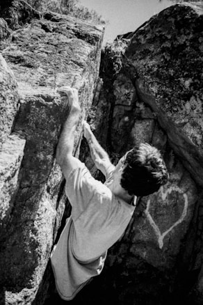 A person climbs a rock face with their hands gripping the stone tightly. The rock surface is rough and weathered with a light-colored heart symbol etched onto it. The person wears a casual t-shirt, and the scene is captured in black and white, giving it a timeless, classic feel.