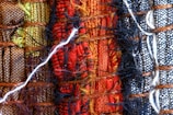 A close-up view of woven textiles featuring a variety of vibrant colors and textures. The arrangement includes sections of brown, green, red, black, and white fabrics, intricately interwoven with visible threads and fibers, including a prominent white thread crossing horizontally.