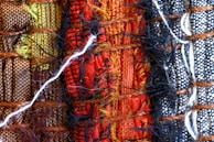 A close-up view of woven textiles featuring a variety of vibrant colors and textures. The arrangement includes sections of brown, green, red, black, and white fabrics, intricately interwoven with visible threads and fibers, including a prominent white thread crossing horizontally.