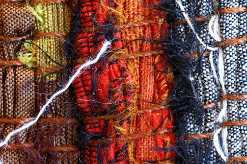 A close-up view of woven textiles featuring a variety of vibrant colors and textures. The arrangement includes sections of brown, green, red, black, and white fabrics, intricately interwoven with visible threads and fibers, including a prominent white thread crossing horizontally.