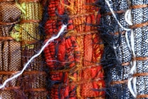 A close-up view of woven textiles featuring a variety of vibrant colors and textures. The arrangement includes sections of brown, green, red, black, and white fabrics, intricately interwoven with visible threads and fibers, including a prominent white thread crossing horizontally.