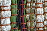 A close-up view of intricately woven fabric patterns featuring a combination of threads in green, beige, and black colors. Thick brown ropes are used to bind sections of the textured fabric, creating an organized yet varied pattern with a distinct tactile quality.