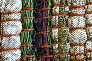 A close-up view of intricately woven fabric patterns featuring a combination of threads in green, beige, and black colors. Thick brown ropes are used to bind sections of the textured fabric, creating an organized yet varied pattern with a distinct tactile quality.