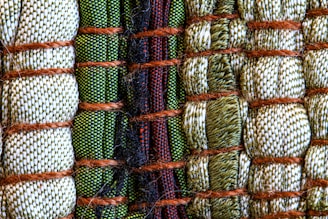 A close-up view of intricately woven fabric patterns featuring a combination of threads in green, beige, and black colors. Thick brown ropes are used to bind sections of the textured fabric, creating an organized yet varied pattern with a distinct tactile quality.