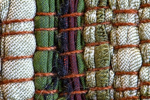 A close-up view of intricately woven fabric patterns featuring a combination of threads in green, beige, and black colors. Thick brown ropes are used to bind sections of the textured fabric, creating an organized yet varied pattern with a distinct tactile quality.