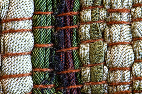 A close-up view of intricately woven fabric patterns featuring a combination of threads in green, beige, and black colors. Thick brown ropes are used to bind sections of the textured fabric, creating an organized yet varied pattern with a distinct tactile quality.