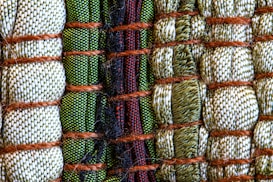 A close-up view of intricately woven fabric patterns featuring a combination of threads in green, beige, and black colors. Thick brown ropes are used to bind sections of the textured fabric, creating an organized yet varied pattern with a distinct tactile quality.