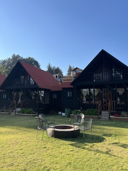 Two rustic wooden cabins with triangular roofs set against a clear blue sky and surrounded by trees. In the foreground, a small grassy lawn features a circular fire pit with four metal chairs arranged around it. The cabins have dark exteriors with red roofs, and are accented with decorative plants near the entrances.