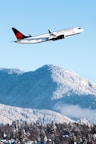 A commercial airplane with the Air Canada logo is flying above snow-covered mountains and a forest, under a clear blue sky. The aircraft is in sharp focus while the mountainous terrain and forest below are blanketed with snow, creating a striking contrast between the white snow and the dark green of the trees.