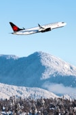 A commercial airplane with the Air Canada logo is flying above snow-covered mountains and a forest, under a clear blue sky. The aircraft is in sharp focus while the mountainous terrain and forest below are blanketed with snow, creating a striking contrast between the white snow and the dark green of the trees.