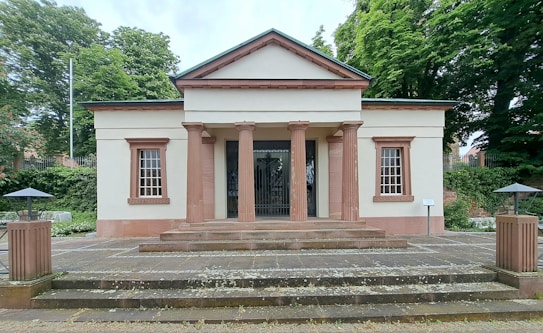 A neoclassical building with a symmetrical facade featuring four columns supporting a triangular pediment. The structure has large windows framed by decorative patterns. It is surrounded by a lush green environment with trees and neatly arranged paved pathways leading to the entrance.