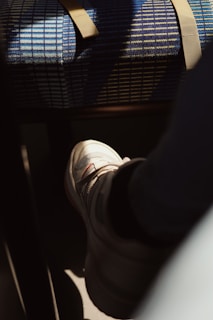 A person wearing a white sneaker with red details. The foot is resting on a surface, possibly a floor, illuminated by a warm, natural light casting shadows. Nearby, there is a blue, grid-patterned fabric with a tan strap draped across it, likely part of a chair or seat.